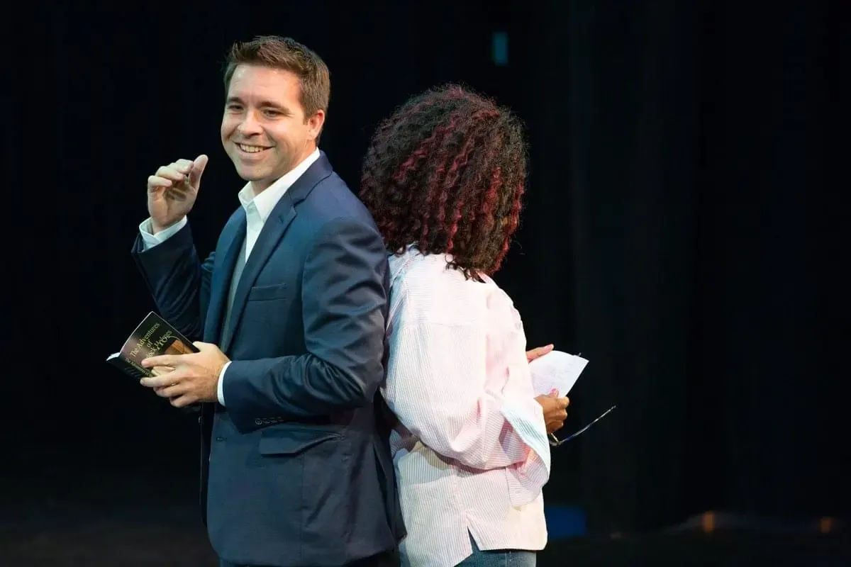 Matt smiling at the camera, standing back-to-back with a guest on a dark stage while holding a small book.