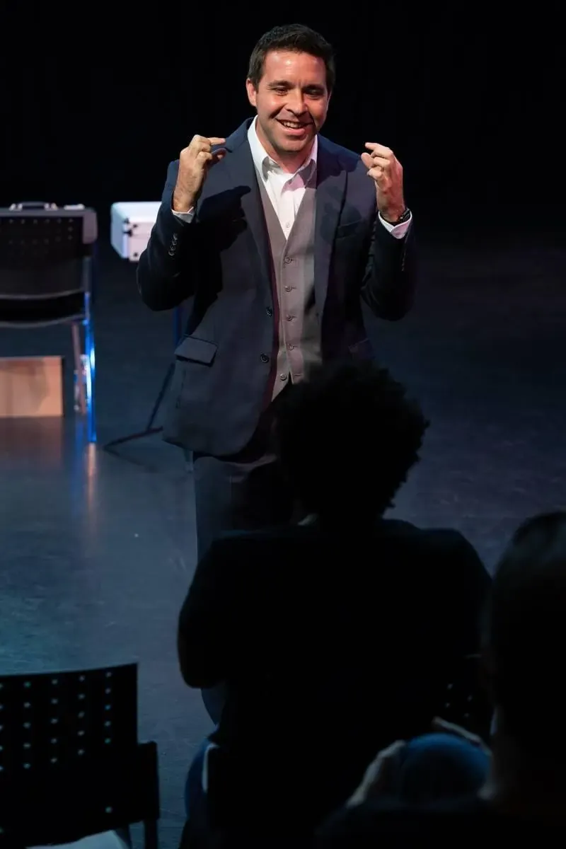 Matt on a darkened stage holding a cup above a silhouetted audience