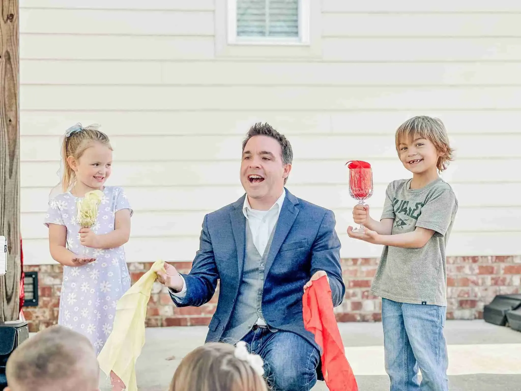 Matt kneeling between two young children on a driveway, one holding a silk, one holding a goblet