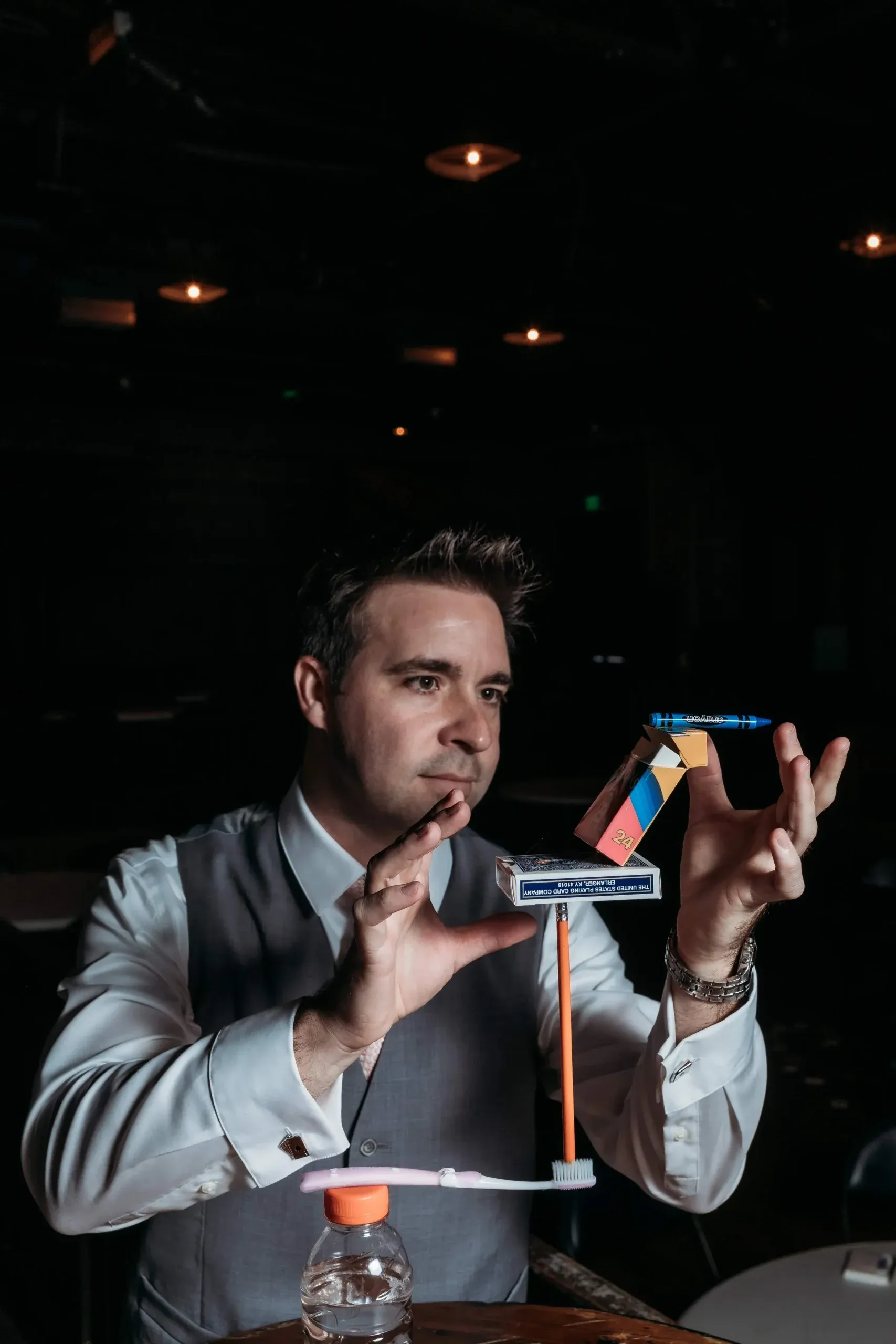 Matt in a waistcoat, balancing a crayon, a card box and a pencil on a toothbrush laid across a water bottle