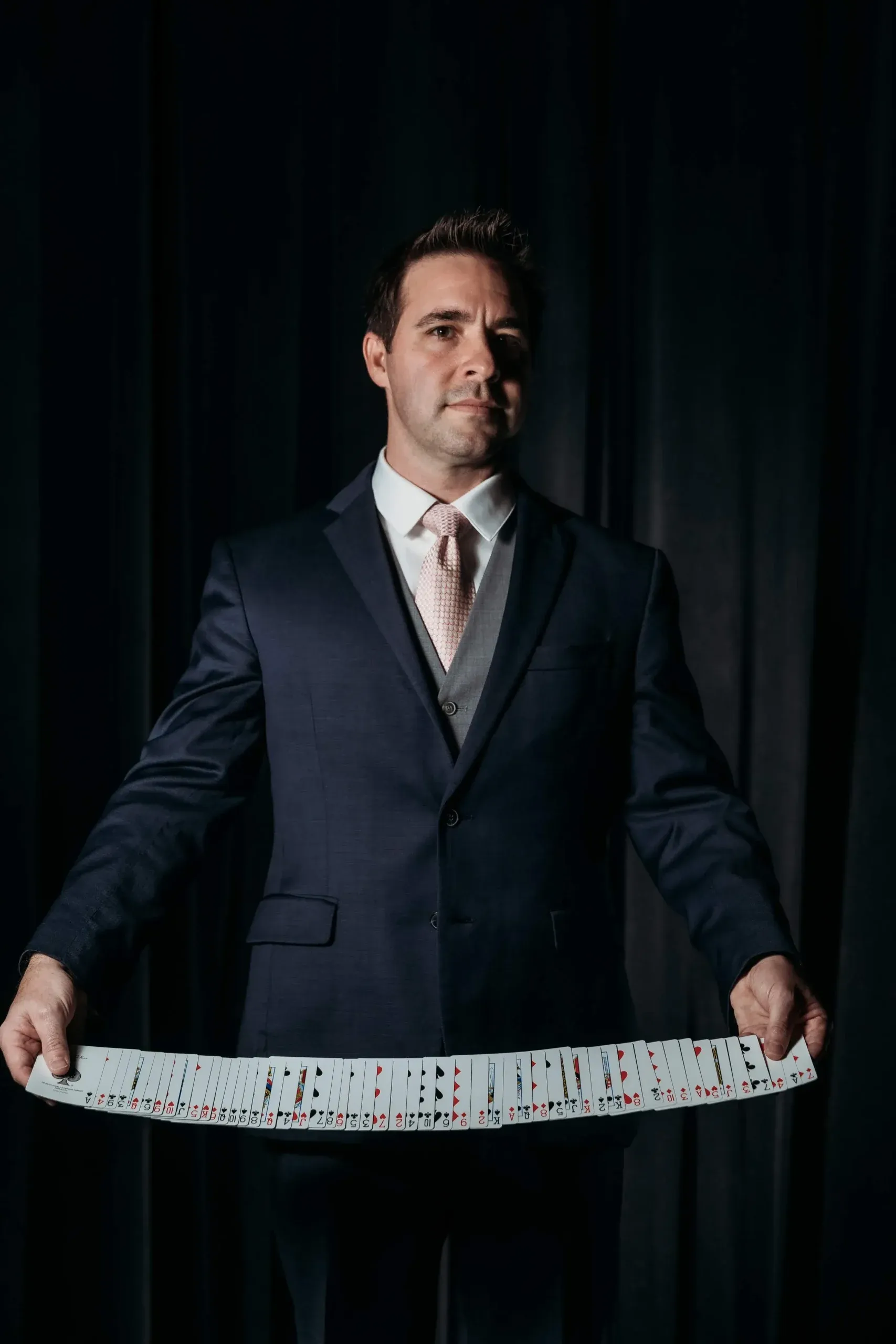Matt Wilson in a dark suit, fanning playing cards in front of a dark curtain backdrop.