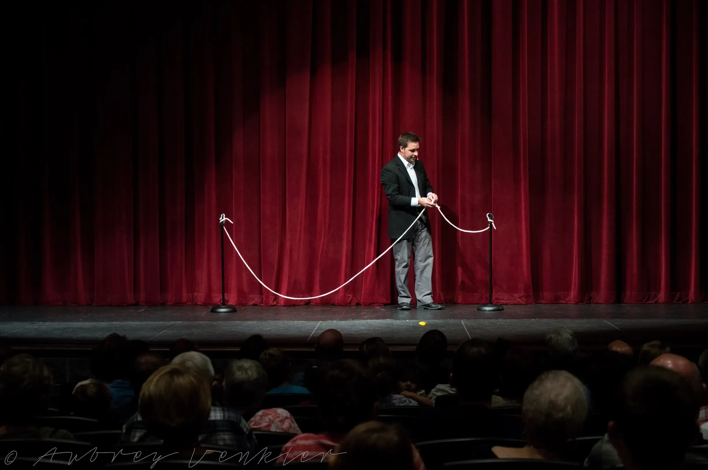 Matt performing a rope routine on a theater stage framed by a red velvet curtain, with rows of the seated audience visible in silhouette below